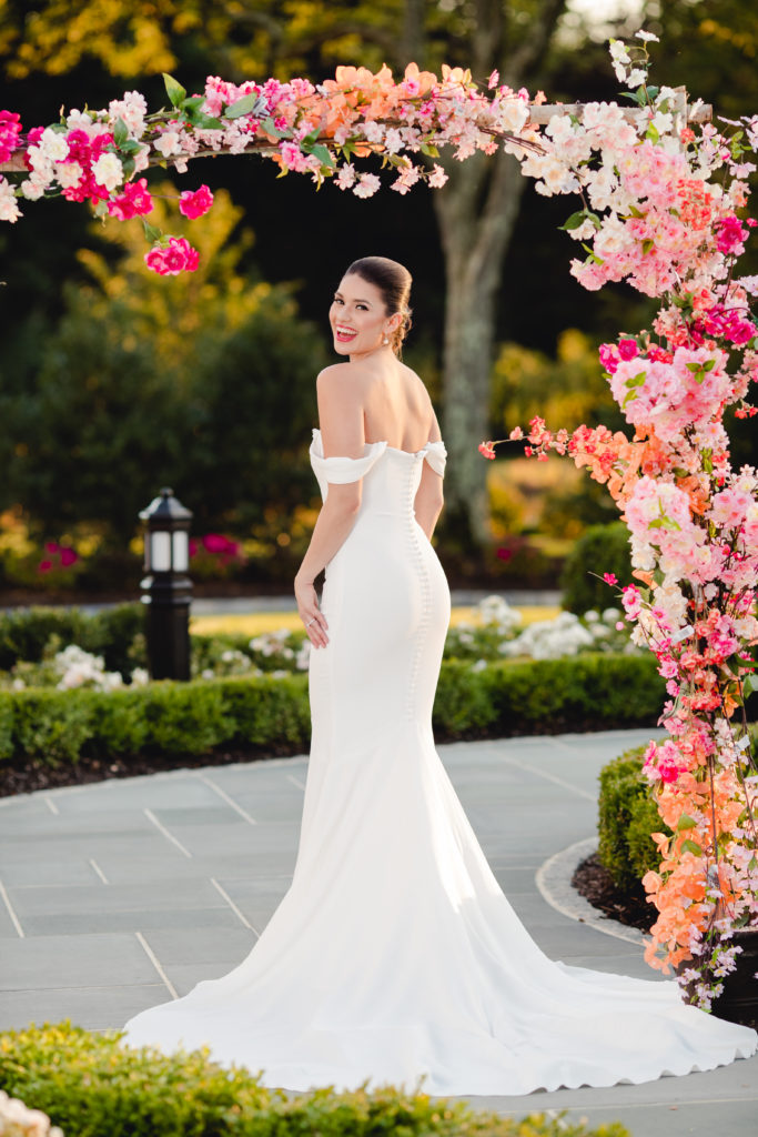 stunning bride in off the shoulder wedding dress posing by pink and white flowers garden. Photo by Vanessa Joy photography