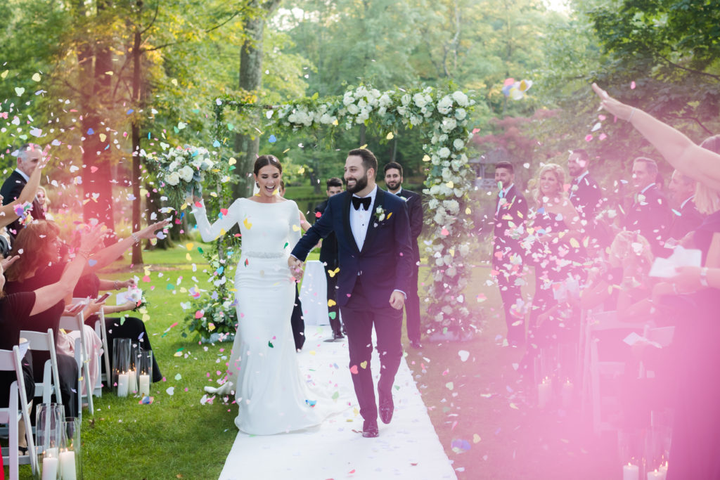 Just Married bride and groom walking down the aisle with family throwing confetti in garden outdoor ceremony. Photo by Vanessa Joy Photography
